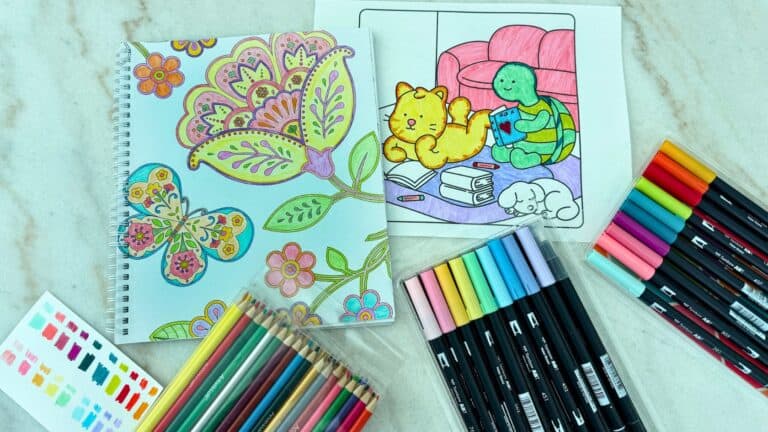 A flatlay of adult coloring pages and a book with colored pencils and markers on a desk