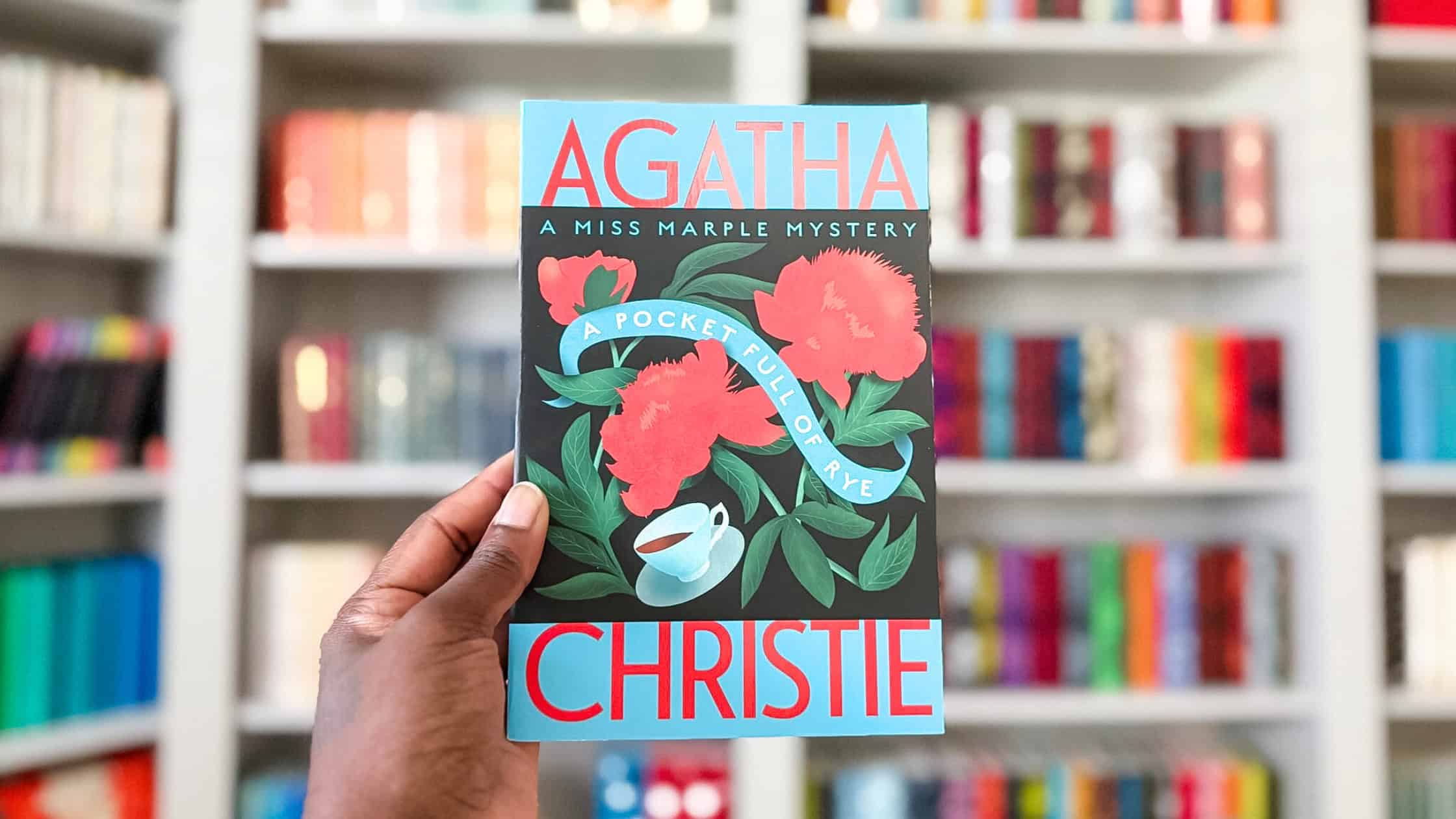 Holding a copy of A Pocket Full of Rye by Agatha Christie with the backdrop of my bookshelves