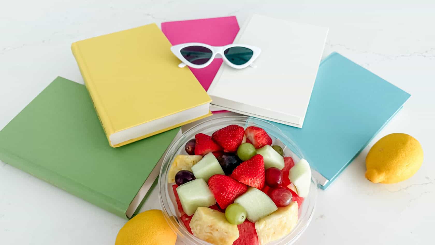 books and sunglasses flatlay with lemons and a bowl of fresh fruit for the summer reading challenge 2026
