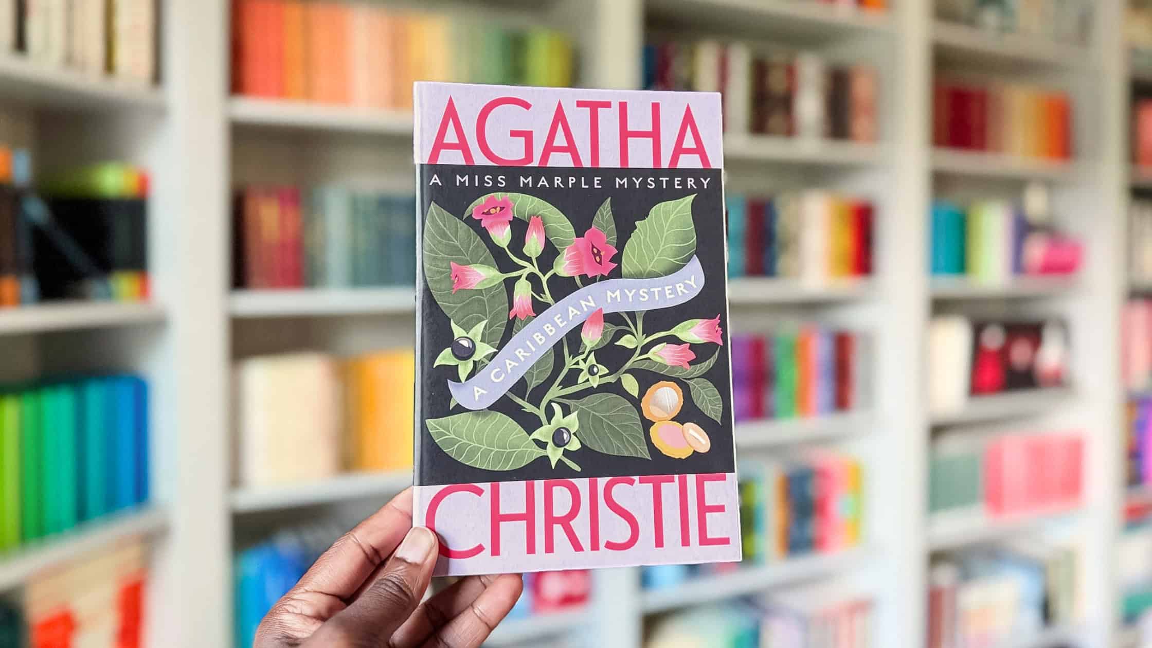 Holding a copy of A Caribbean Mystery by Agatha Christie with the backdrop of my bookshelves